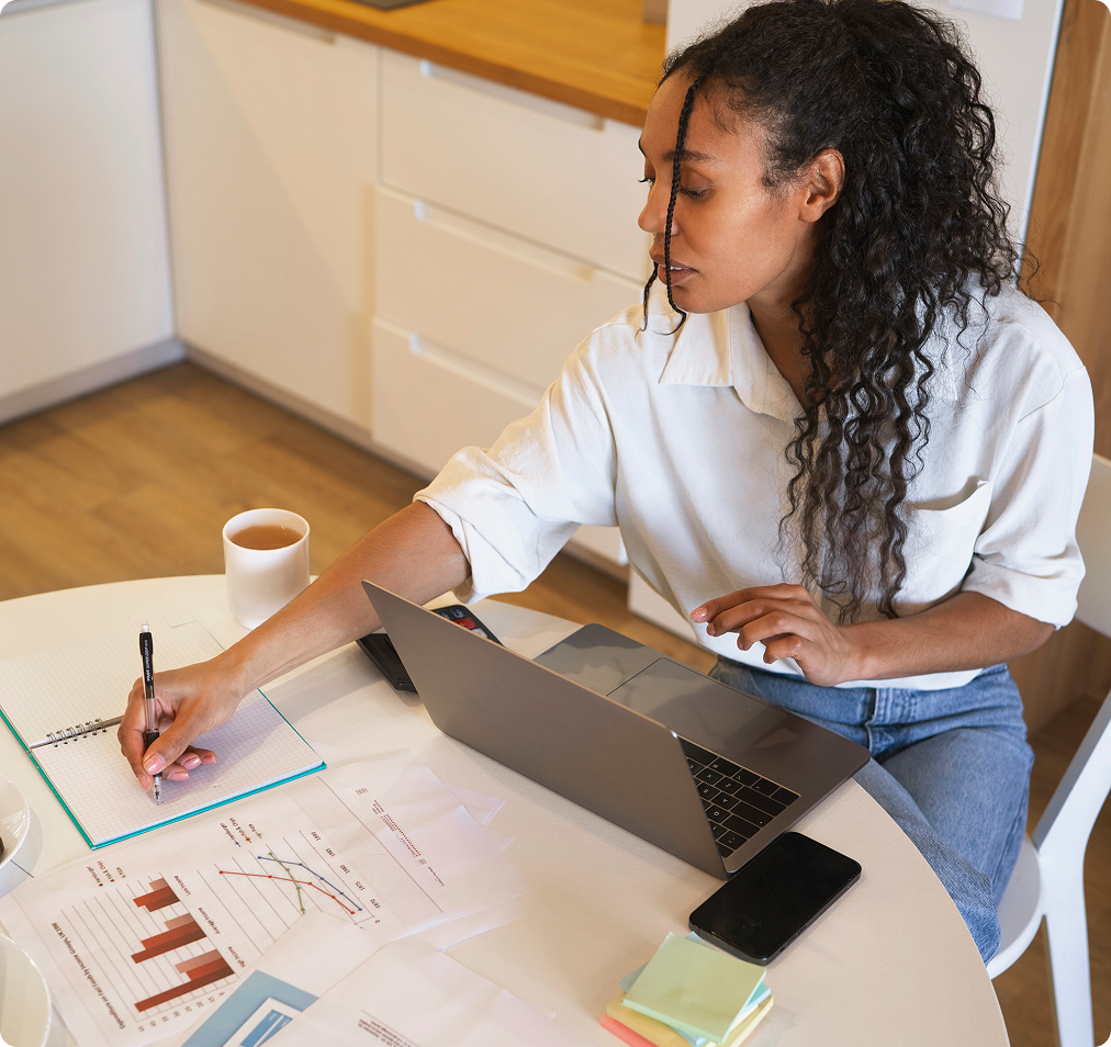 A person with a laptop taking notes, with a messy desk full of sticky notes, charts, and paper.